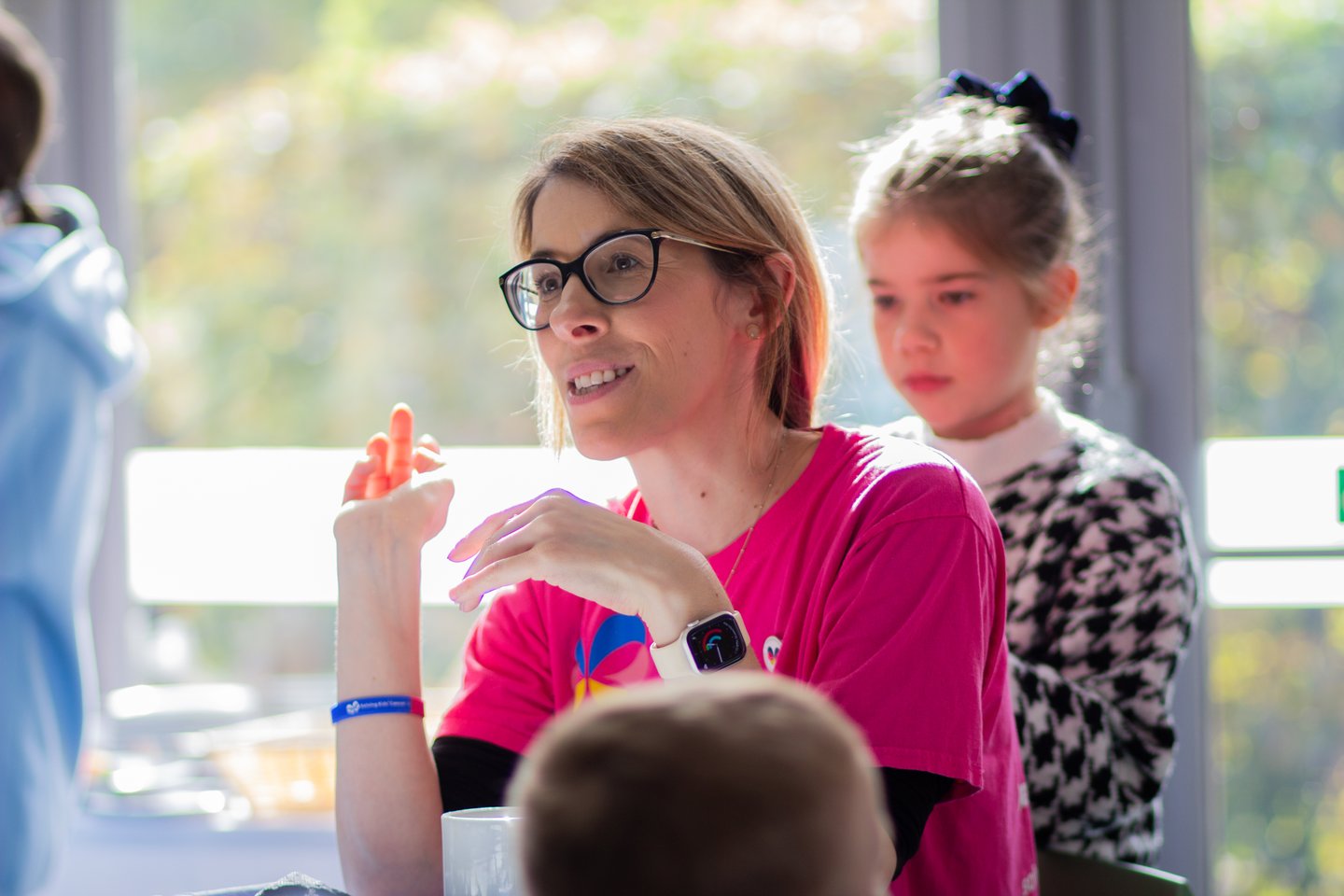 Person wearing a bright pink Solving Kids’ Cancer UK t-shirt seated at a table during a family event, speaking with other attendees.