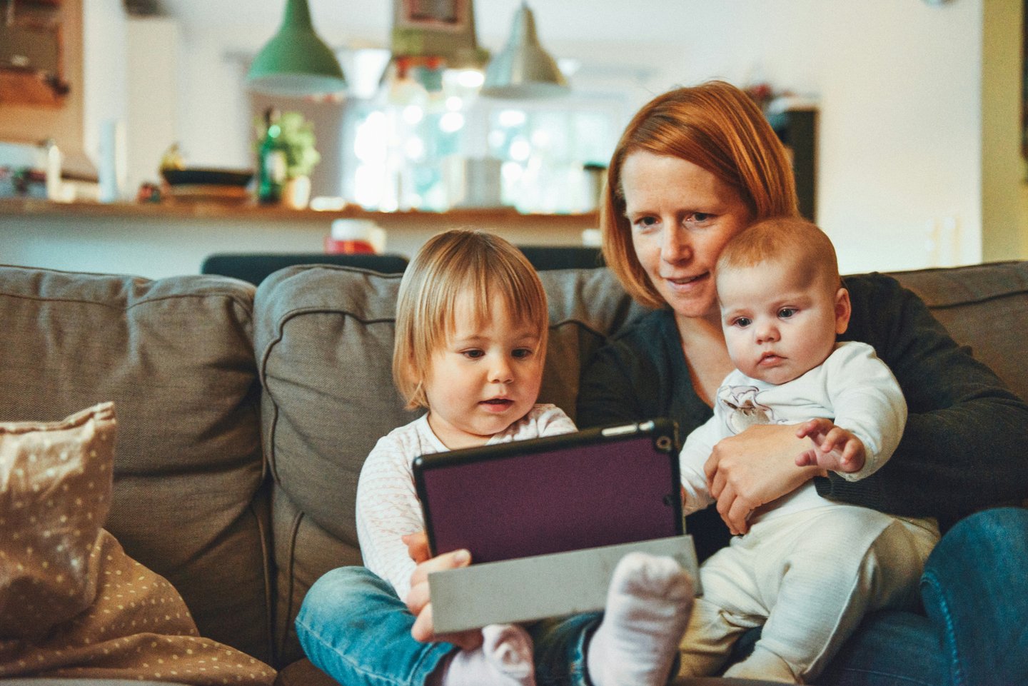 Mother with a baby and toddler sitting on a sofa and looking at a tablet together.