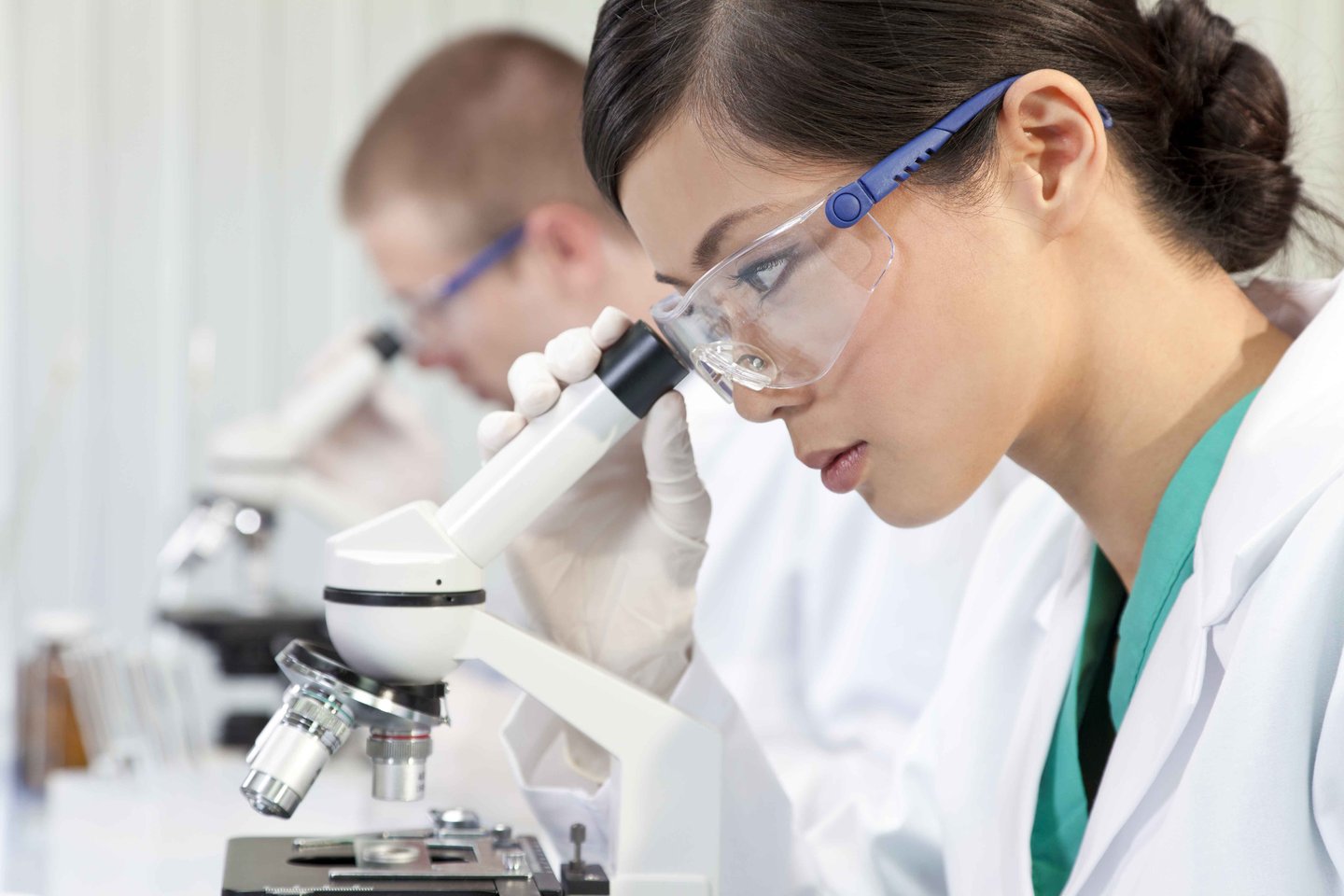 A scientist, wearing lab goggles, gloves and coat, looks down a white microscope. Another scientist is doing the same, in an out of focus background.