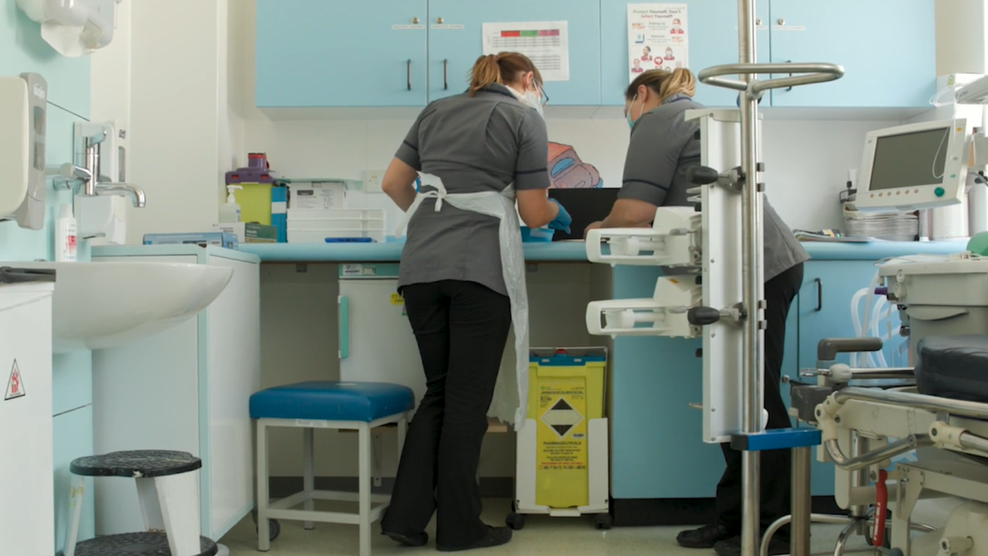 Two nurses in uniforms and masks work together at a countertop in a clinical room, with medical equipment visible around them.