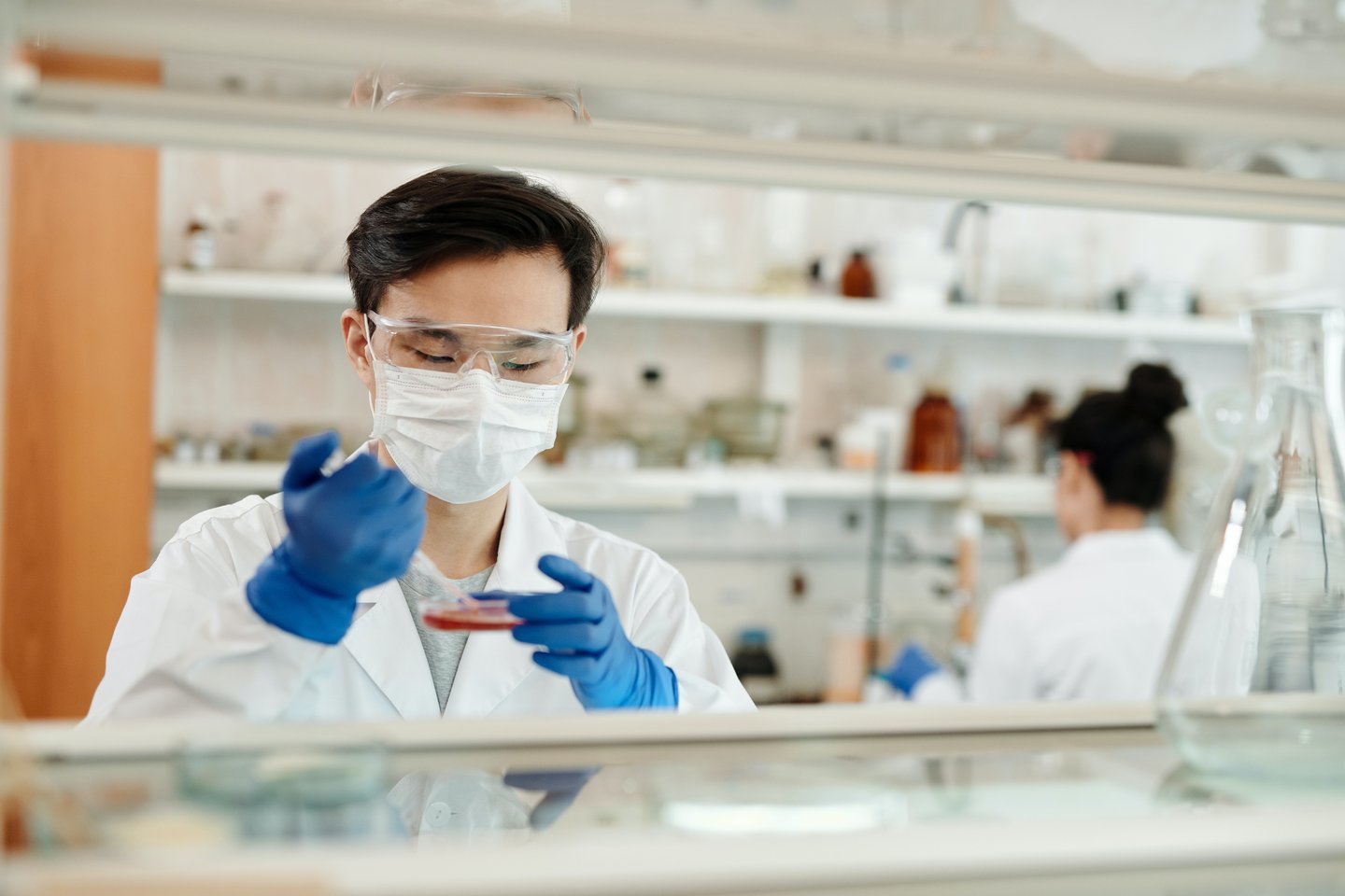 Scientist wearing protective goggles, a face mask, and blue gloves works with a petri dish in a lab, with equipment and a colleague in the background.