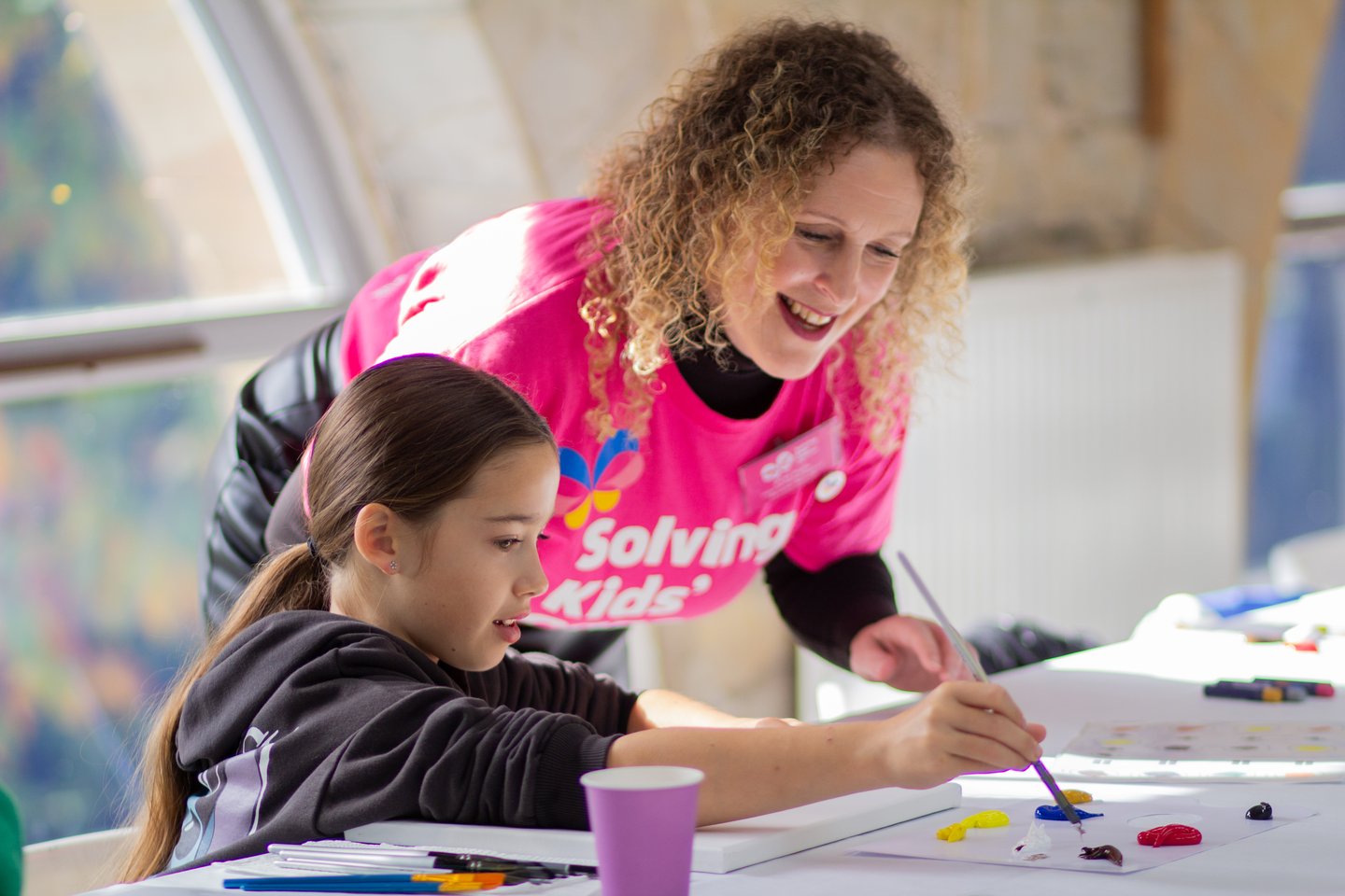 Child painting at a table during a creative activity, with a member of Solving Kids’ Cancer UK staff providing support.