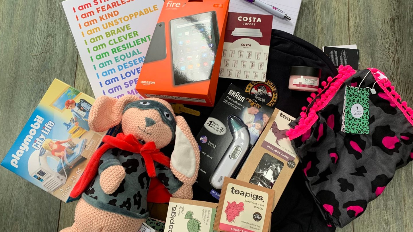 Contents of a family support package laid out on a table, including a motivational notebook, Amazon Fire tablet, soft toy rabbit, digital thermometer, tea bags, Costa Coffee voucher card and stationery items.