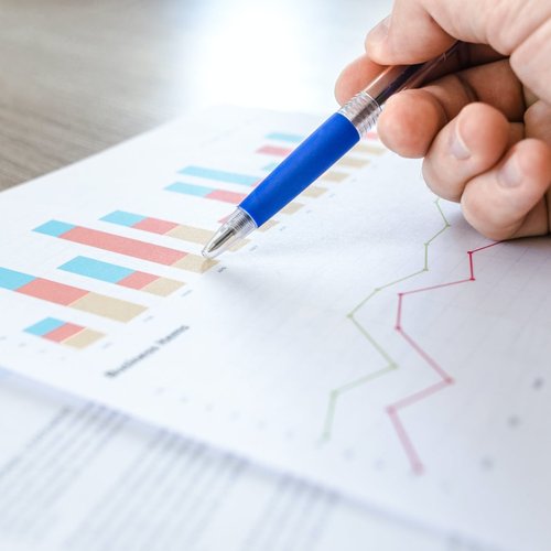 A hand holding a blue pen rests on documents displaying a colourful bar chart and a line chart, suggesting statistical analysis.