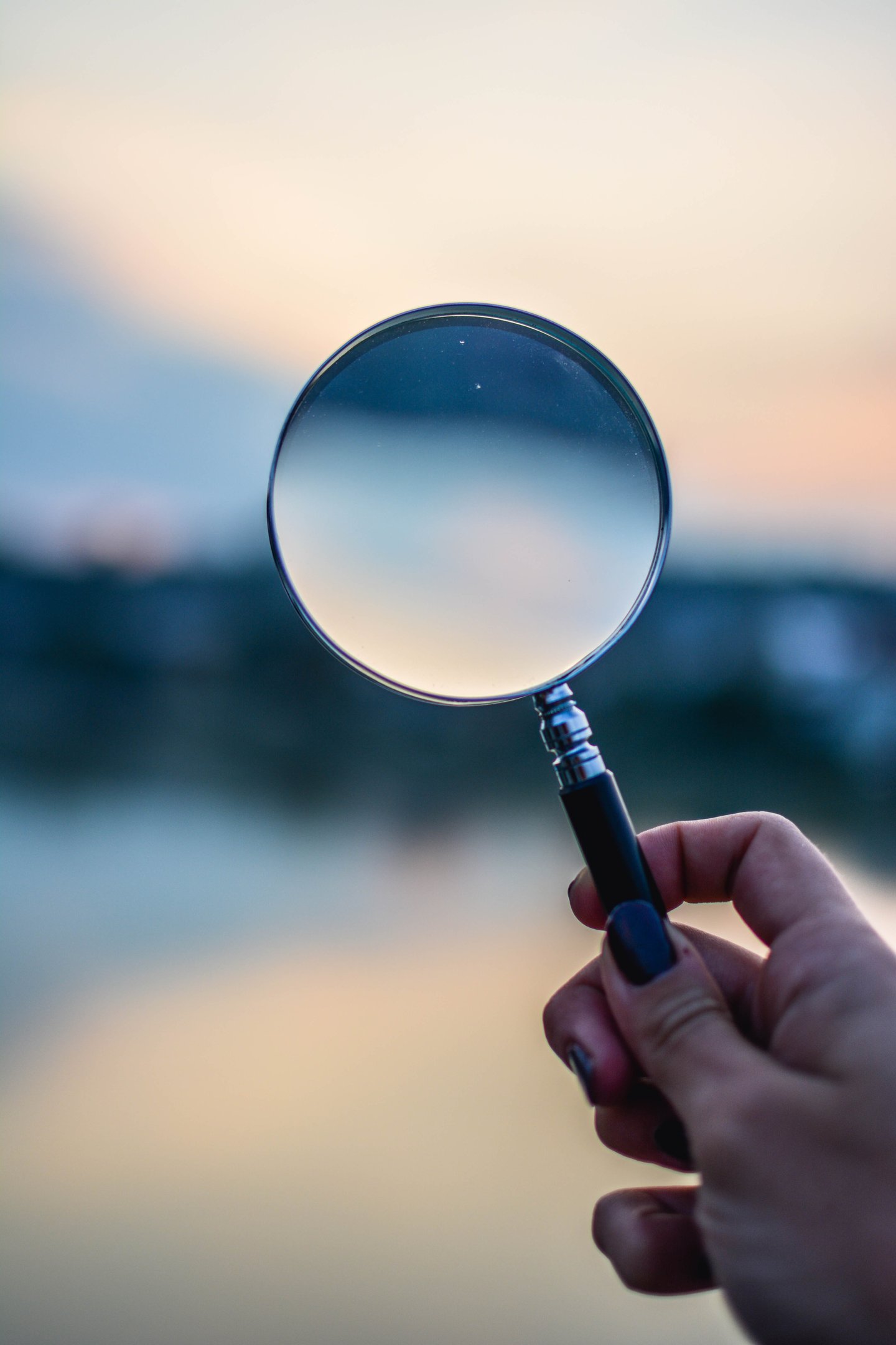 A hand holding-up a magnifying glass against an out of focus blue skyline.