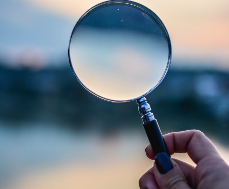A hand holding-up a magnifying glass against an out of focus blue skyline.