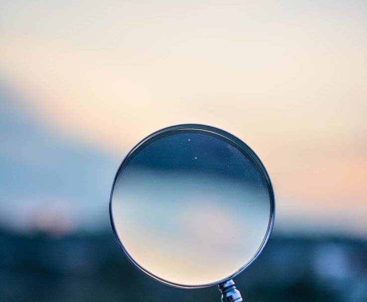 A hand holding-up a magnifying glass against an out of focus blue skyline.