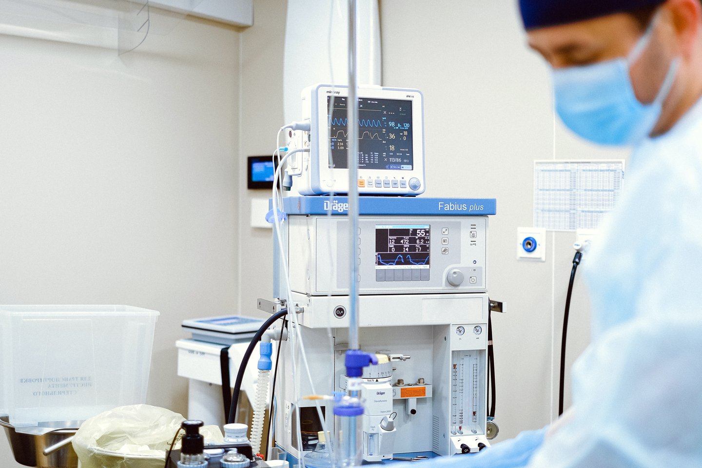 Medical machine with a screen showing patient data, IV lines in front, and a masked healthcare worker in the background.