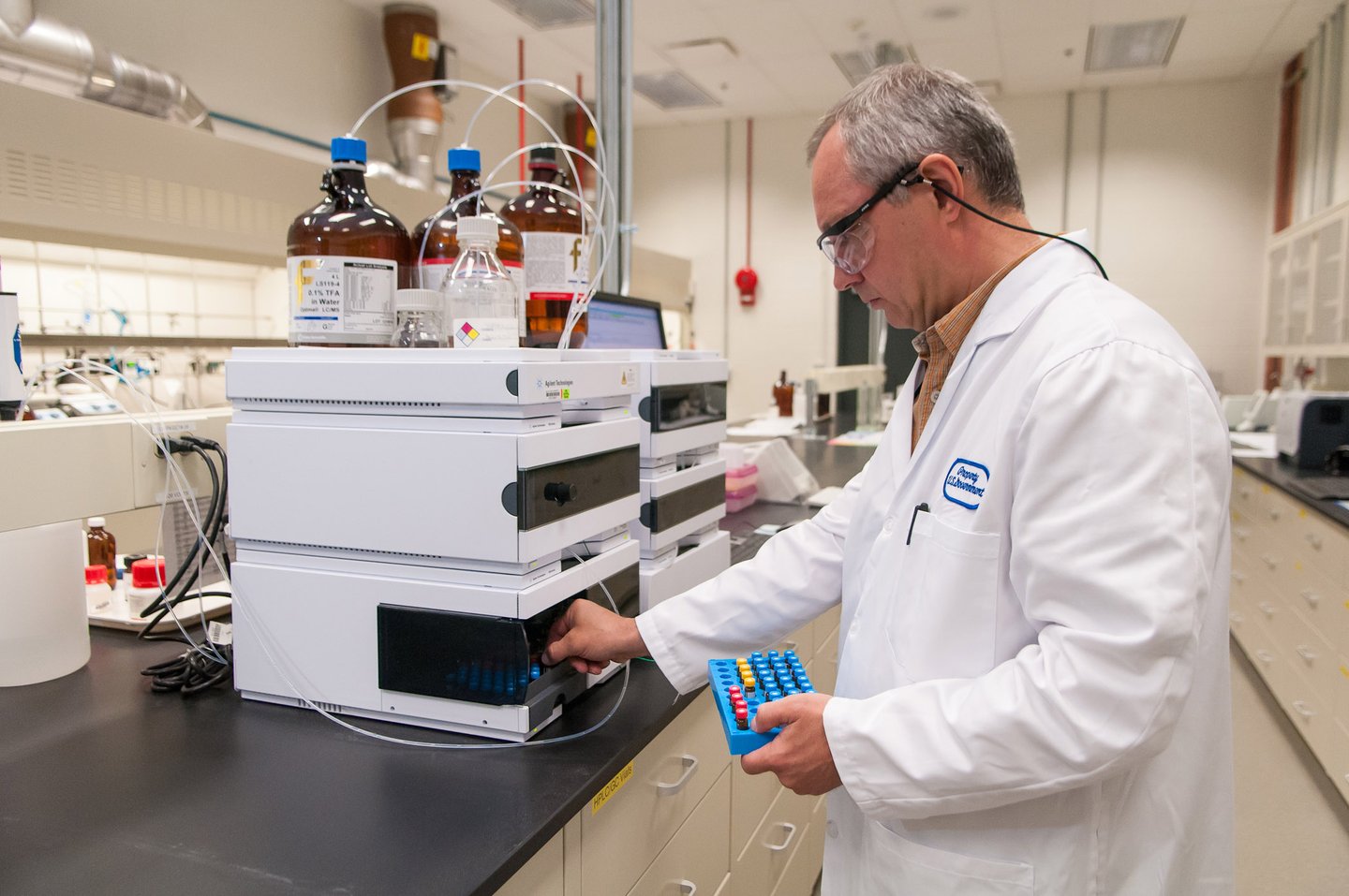 A scientist in a lab coat and safety goggles operates advanced lab equipment, holding a tray of vials, in a well-equipped research laboratory.