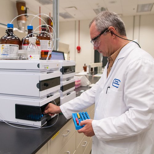 A scientist in a lab coat and safety goggles operates advanced lab equipment, holding a tray of vials, in a well-equipped research laboratory.