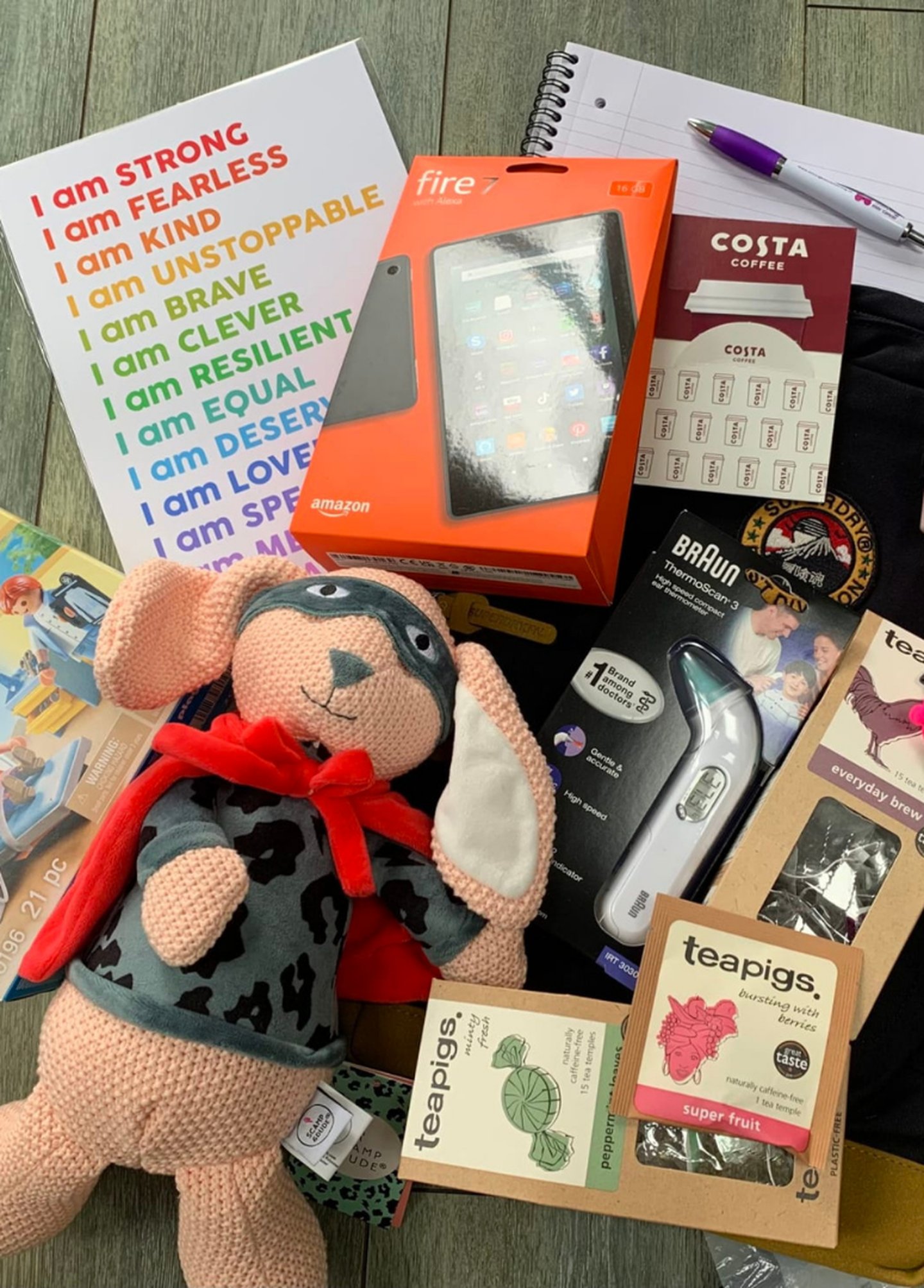 Contents of a family support package laid out on a table, including a motivational notebook, Amazon Fire tablet, soft toy rabbit, digital thermometer, tea bags, Costa Coffee voucher card and stationery items.