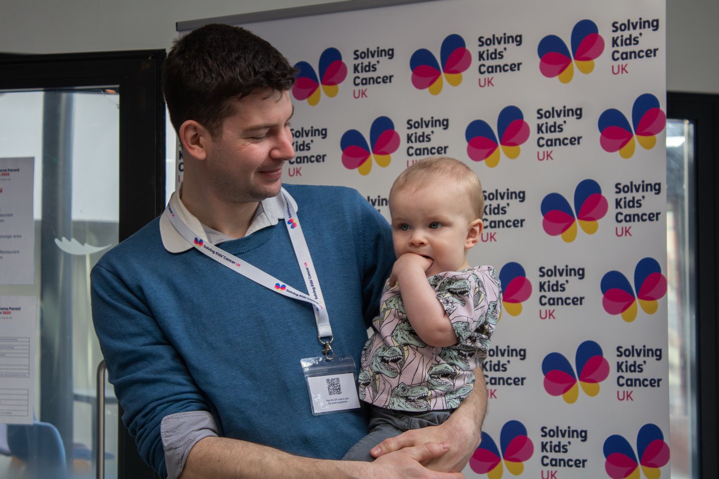 Picture of baby boy with his Dad in front of a Solving Kids' Cancer UK branded banner