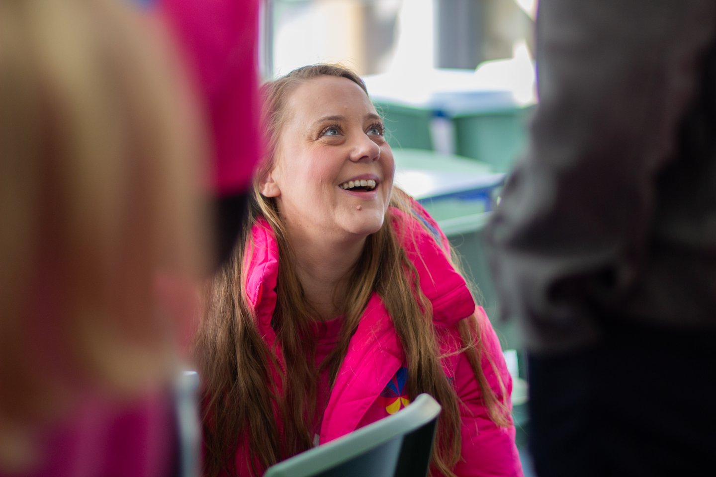 Person wearing a bright pink Solving Kids’ Cancer UK jacket seated indoors at a family event, looking up while engaging in conversation.