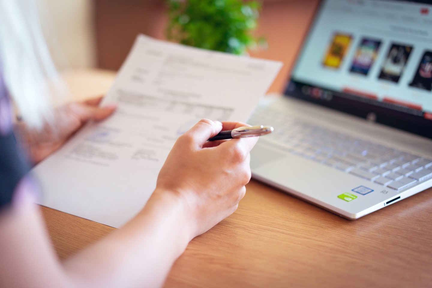 A close-up of a person's hands holding a printed document in one hand and a pen in the other, at a desk with an open laptop in the background.