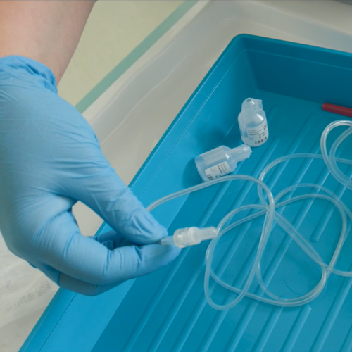 A close-up of a gloved hand holding clear medical tubing connected to a syringe, placed inside a blue sterile tray with two small containers inside.