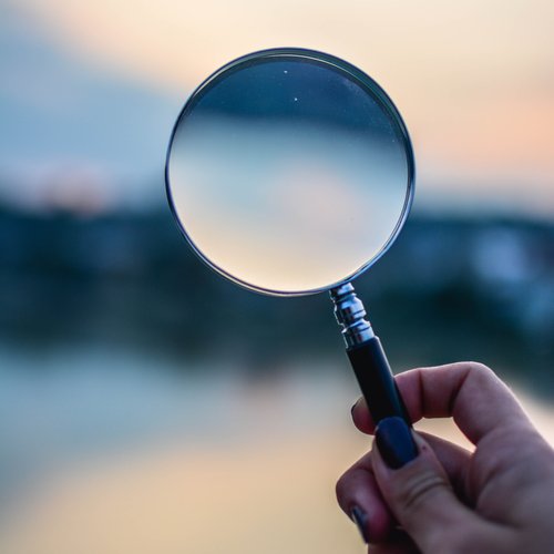 A hand holding-up a magnifying glass against an out of focus blue skyline.