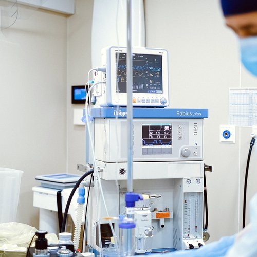 Medical machine with a screen showing patient data, IV lines in front, and a masked healthcare worker in the background.