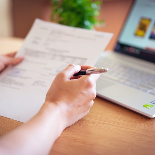 A close-up of a person's hands holding a printed document in one hand and a pen in the other, at a desk with an open laptop in the background.